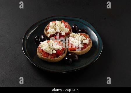 Nahansicht der kretischen Dakos auf dem Teller, bestehend aus gerstenrusk mit saftigen Tomaten, Fetakäse und Olivenöl auf dunklem Hintergrund. Stockfoto