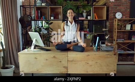 Frau, lächelnde Büroangestellte, die auf dem Schreibtisch sitzt, umgeben von Computer und Bürobedarf, und beruhigende Meditation in Yoga-Lotus-Pose macht Stockfoto