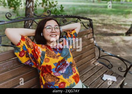 Eine Asiatin genießt ihre Zeit allein auf einer Parkbank, während sich eine lächelnde Frau in der Natur mit entspannten und fröhlichen Melodien entspannt Stockfoto