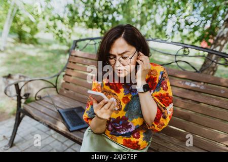 Verwirrte Frau, die auf einer Bank im Park saß, wütend über ihr Telefon sprach, ärgerte sich die junge Dame mit einer Brille, die während des Meeres ein hitziges Telefongespräch führte Stockfoto