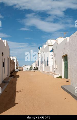 Die Straße ist mit Sand bedeckt. Weiße kleine Häuser auf beiden Seiten. Grüne oder blaue Holztüren und Fensterläden. Blauer Himmel mit weißen Wolken. Caleta del Sebo Stockfoto