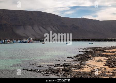 Dunkelbraune bis schwarze Farbe der Klippen im Norden von Lanzarote. Flaches Wasser eines Strandes. Boote in einem Hafen im Hintergrund. Blauer Himmel mit weißem cl Stockfoto