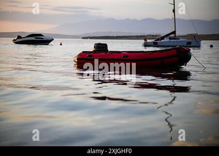 Rotes Schlauchboot schwimmt auf ruhigem Wasser mit Segelbooten und Bergen im Hintergrund. Stockfoto