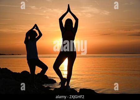 Mutter und Tochter üben Yoga am Strand bei Sonnenuntergang und genießen einen friedlichen Moment zusammen. Stockfoto