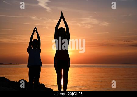 Mutter und Tochter üben Yoga am Strand bei Sonnenuntergang und genießen einen friedlichen Moment zusammen. Stockfoto