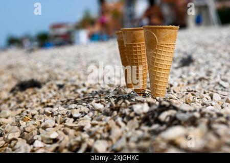 Genießen Sie die Sommerstimmung mit leeren Eiswaffelkegeln an einem Kiesstrand, vor dem glitzernden Meer als Kulisse. Stockfoto