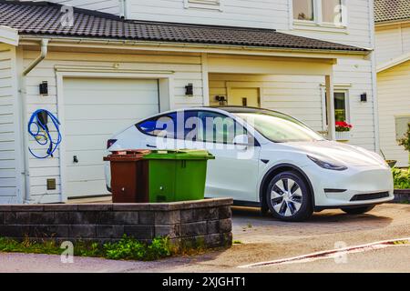 Blick auf das weiße Elektroauto, das vor dem modernen weißen Haus mit Garage und Mülltonnen in der Nähe der Einfahrt geparkt ist. Schweden. Stockfoto