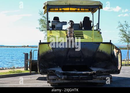 Gelber Asphaltfertiger mit leerem Ladeschaufel, der an einem sonnigen Tag auf einer Straße in der Nähe des Meeres steht. Stockfoto