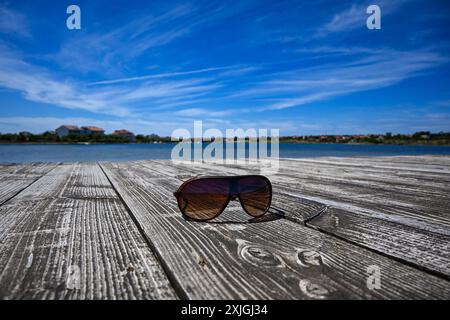 Sonnenbrillen reflektieren den Blick auf das Meer, während sie an einem sonnigen Sommertag auf einer Holzterrasse liegen. Stockfoto