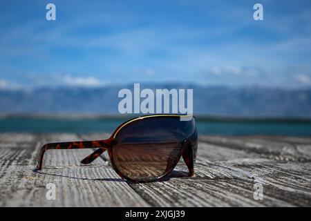 Sonnenbrillen reflektieren den Blick auf das Meer, während sie an einem sonnigen Sommertag auf einer Holzterrasse liegen. Stockfoto