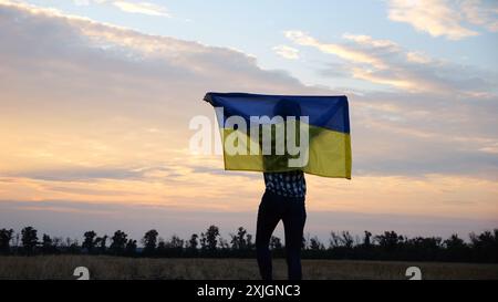 Ukrainische Frau, die mit gehisster Flagge der Ukraine über Kopf auf Weizenfeld vor dem Hintergrund des schönen Sonnenuntergangs geht. Dame, die mit nationalem Banner läuft Stockfoto