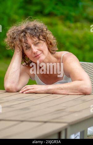 Porträt einer romantischen älteren Frau 50-55 Jahre alt mit lockigen Haaren an einem Tisch im Hintergrund eines Sommergartens Stockfoto