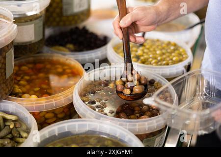 Die Hände der Herren halten die Kelle und legen eingelegte schwarze Oliven mit Stein in einen Einwegbehälter aus Kunststoff Stockfoto