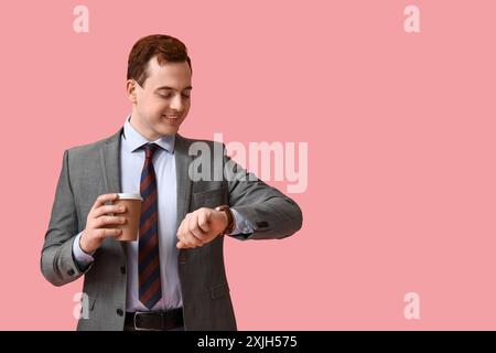 Hübscher junger Mann mit einer Tasse frischen Kaffees, der auf Uhren auf rosa Hintergrund blickt Stockfoto