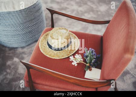 Ein Strohhut mit schwarzem Band, ein kleiner Blumenstrauß aus lila und weißen Blumen und ein leerer Umschlag ruhen auf einem roten Polsterstuhl. Ein blau gestreifter Hocker Stockfoto