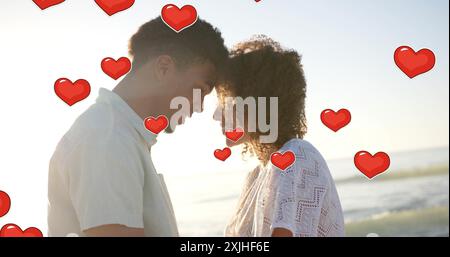 Bild von Herzen, die sich über verschiedene verliebte Paare bewegen, die im Sommer Köpfe am Strand berühren Stockfoto