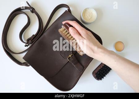 Fotoverfahren zur Reinigung von Lederprodukten. Ledertasche mit verrücktem Pferd. Menschliche Hand mit einem Schwamm. Bürsten und Wachs sind in der Nähe. Die Tasche ist braun. Weißer Hintergrund. Stockfoto