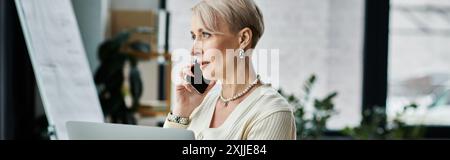 Geschäftsfrau mittleren Alters mit kurzen Haaren, die in einem modernen Büro mit einem Handy spricht und einen Laptop hält. Stockfoto