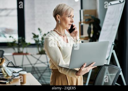 Eine Geschäftsfrau mittleren Alters mit kurzen Haaren spricht auf einem Handy, während sie in einem modernen Büro einen Laptop hält. Stockfoto