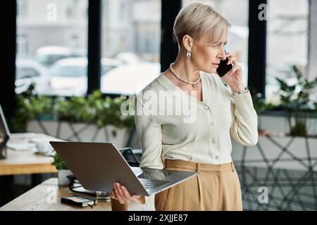Eine Geschäftsfrau mittleren Alters mit kurzen Haaren, die in einem modernen Büro mit einem Handy spricht und einen Laptop hält. Stockfoto