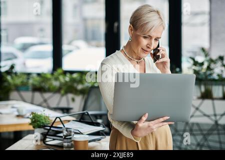 Geschäftsfrau mittleren Alters mit kurzen Haaren, die in einem modernen Büroumfeld auf dem Handy spricht, während sie auf dem Laptop schreibt. Stockfoto