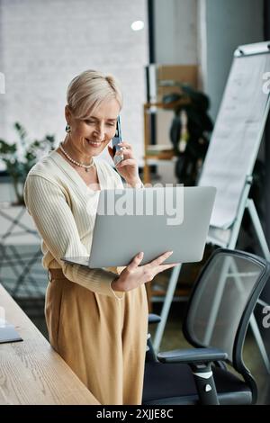 Eine Geschäftsfrau mittleren Alters mit kurzen Haaren spricht gleichzeitig auf einem Handy und arbeitet in einem modernen Büro an einem Laptop. Stockfoto