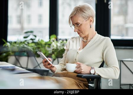 Geschäftsfrau mittleren Alters mit kurzen Haaren, die auf einem Stuhl sitzt und sich in einem modernen Büro auf das Mobiltelefon konzentriert. Stockfoto