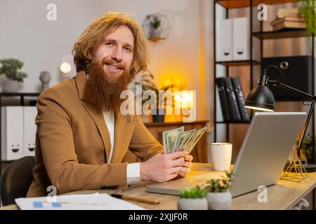 Glücklicher kaukasischer Geschäftsmann mit rotem Haar, der Geld in Bargeld zählt und im Heimbüro einen Laptop benutzt. Ein erfolgreicher Freelancer, der Geld im Amt bekommt, berechnet Dividenden Geld Geld Gewinne Finanzen. Stockfoto