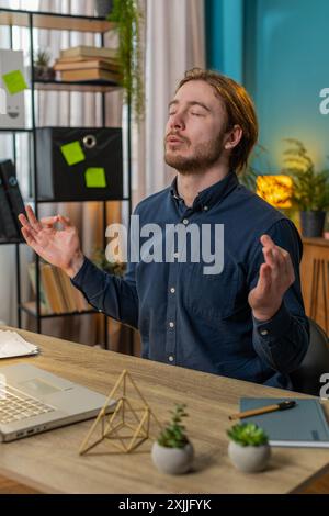 Friedlicher, entspannter männlicher Geschäftsmann, der am Arbeitsplatz mit gefalteten Mudra-Händen sitzt, Stress während des Arbeitstages reduziert, meditiert oder Yoga-Atemübungen im Heimbüro macht. Vertikal Stockfoto
