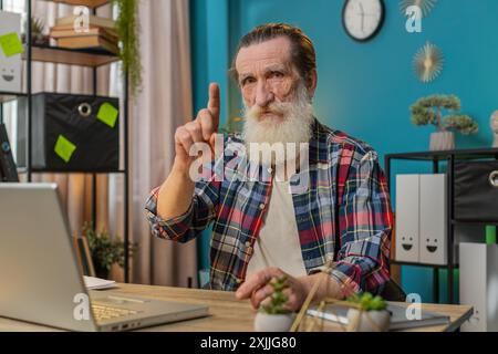 Kaukasischer Senior Mann, der an einem Laptop arbeitet, schüttelt den Finger und sagt Nein. Seien Sie vorsichtig, schimpfen Sie und geben Sie Ratschläge, um Gefahren-Fehler-Missbilligung-Schild im Home Office zu vermeiden. Freiberuflicher Großvater sitzt am Schreibtisch Stockfoto