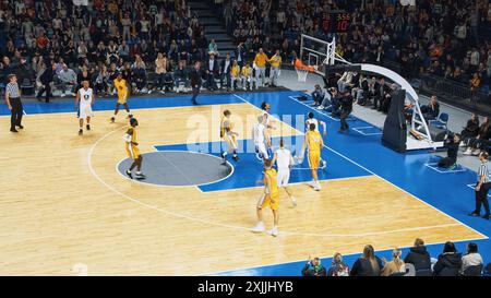 Basketball-Meisterschaft in einer überfüllten Arena: Gelbes Team, das den Ball zwischen seinen Teamkollegen durchführt, wobei der Forward aus der Entfernung ein zwei-Punkte-Tor erzielt. Sport TV Channel Broadcast Konzept. Stockfoto