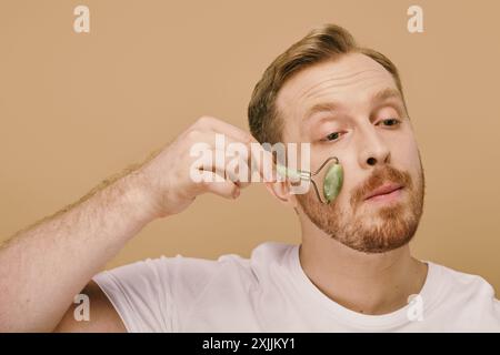 Ein Mann in lässiger Kleidung, der sanft Gesichtsroller für eine Hautpflegeroutine benutzt. Stockfoto