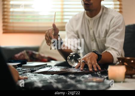 Lächelnder männlicher Wahrsager liest Tarotkarte am Tisch mit brennenden Kerzen und Kristallkugel Stockfoto