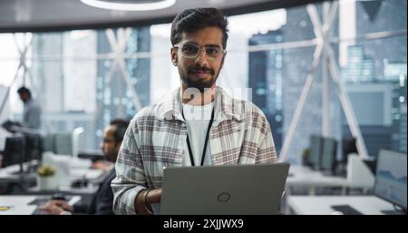 Hübscher indischer Mann mit Brille, charmant lächelnd und in die Kamera schaut. Junger intelligenter männlicher Ingenieur, der einen Laptop hält. Finance Manager, der in modernen Unternehmensbüros arbeitet Stockfoto