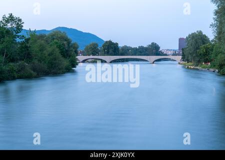 Der Fluss fliegt in der Nähe der Stadt. Abendblick mit eingeschalteten ersten Lichtern. Langzeitbelichtung mit geglättetem Wasser. Stockfoto