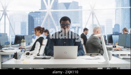 Erfolgreicher Schwarzer Mann, Der Auf Laptop-Computer Im Hellen Scyscraper Office Mit Verschiedenen Mitarbeitern In Downtown Tippt. Cheerful Male Programmer Coding In Modern Fintech Startup Workspace, Engineering Software Stockfoto