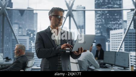 Erfolgreicher Kaukasischer Geschäftsmann, Der Im Büro Mit Verschiedenen Mitarbeitern In Downtown Skyscraper Unterwegs Ist. Unternehmer mittleren Alters mit Laptop im E-Commerce Company Workspace. Dramatischer Abend Bearbeiten. Stockfoto