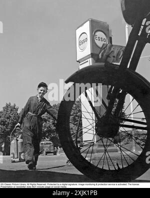 ESSO-Tankstelle in den 1950er Jahren Ein junger Mann sieht serviceorientiert aus, wenn er glücklich auf der Arbeit ist, Autos und Motorräder zu füllen und Öl zu prüfen. Stockfoto