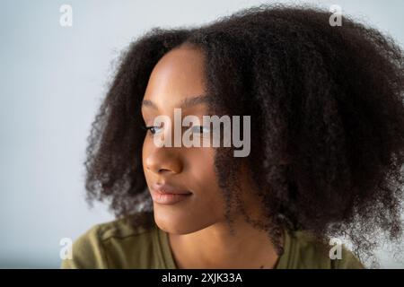 Junge afroamerikanische Frau mit natürlichen lockigen Haaren, die nachdenklich aussieht, vor hellem Hintergrund. Stockfoto