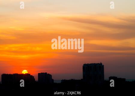 Sonnenaufgang in einem bewölkten Sommerhimmel über der Stadt. Die aufgehende Sonne bricht durch helle Wolken vor dem Hintergrund von Silhouetten von Gebäuden Stockfoto