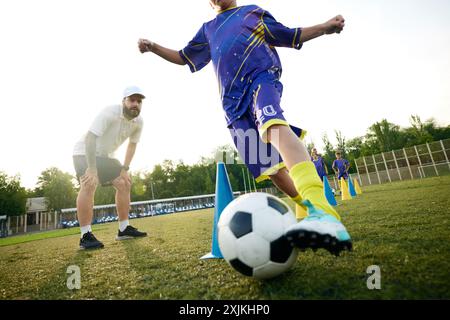 Kleiner Junge, Kind in Bewegung, dribbelt Fußball um Kegel und erhält Anleitung vom Trainer. Stadionspiel im Freien. Konzentriere dich auf Beine und Ball Stockfoto