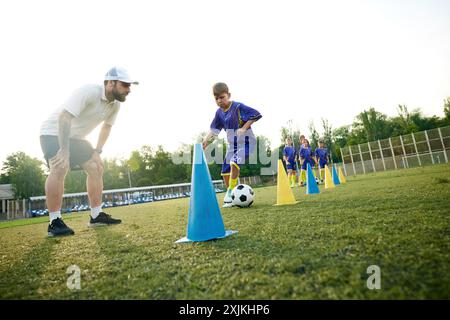 Kleiner Junge, Kind in Bewegung, dribbelt Fußball um Kegel und erhält Anleitung vom Trainer. Stadionspiel im Freien Stockfoto