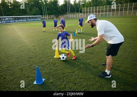 Der Junge übt das Dribbeln durch Kegel mit gezielter Unterstützung durch den Trainer. Stadion im Freien und Spiel am Abend. Stockfoto