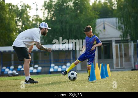 Junge, Kind in lila Uniform in Bewegung, verbessert Dribbling-Fähigkeiten und erhält Anleitung vom Trainer. Outdoor-Training Stockfoto