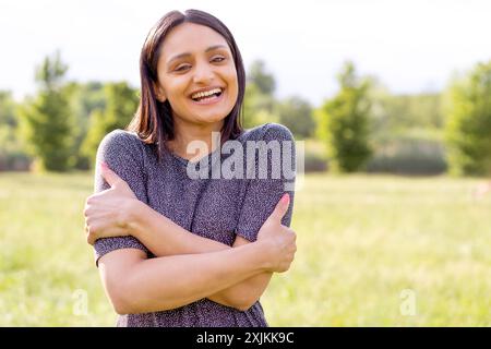 Porträt einer fröhlichen jungen Indianerin mit überkreuzten Armen, lachend in einem sonnigen Park. Stellt Glück, Selbstvertrauen und Wellness in einem natürlichen Set dar Stockfoto