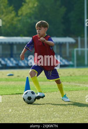 Vertikales Bild von konzentriertem Jungen, Kind in rot und blau Sportswear Dribbling Ball durch das Feld mit Fokus, Training Fußballspiel. Stockfoto