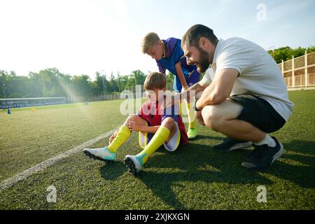 Coach, der Kindern, Jungen, der auf dem Feld sitzt und Schmerzen an den Beinen spürt, während des Spiels verletzt wird. Stockfoto