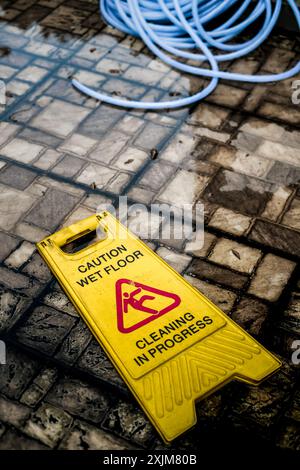 Gelber Hinweis: Reinigung des nassen Fußbodens läuft, Schild und Gartenschlauch fiel auf dreckigen, feuchten und überfluteten Außenboden Stockfoto