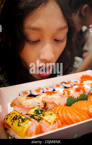 Südostasiatisches Teenager-Mädchen, das eine Kiste Sushi und Sashimi japanisches Essen ansieht, bento-Mahlzeit mit weit geöffnetem Mund Stockfoto