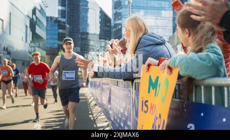 Marathon-Publikum unterstützt und bejubelt ihre Lieben, die am Rennen teilnehmen: Athletic Male Marathon Runner gibt eine High Five an weibliche Familienmitglieder im Publikum während des Laufens Stockfoto